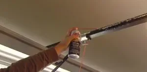 A hand spraying lubricant from an aerosol can onto a garage door opener rail.