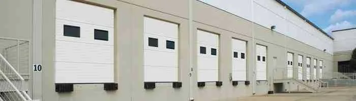 A row of white insulated overhead doors with small windows along the loading docks of a large commercial building.