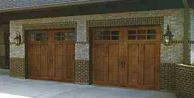 Two wood carriage house doors with square windows on a stone and brick house with coach lights.