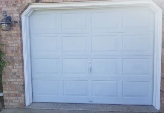 A white raised-panel single garage door with a center handle, in white trim set into a brick wall, with a coach light at the left.