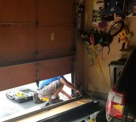 A worker kneels under a partly raised door, working at its bottom section with an open tool case beside him.